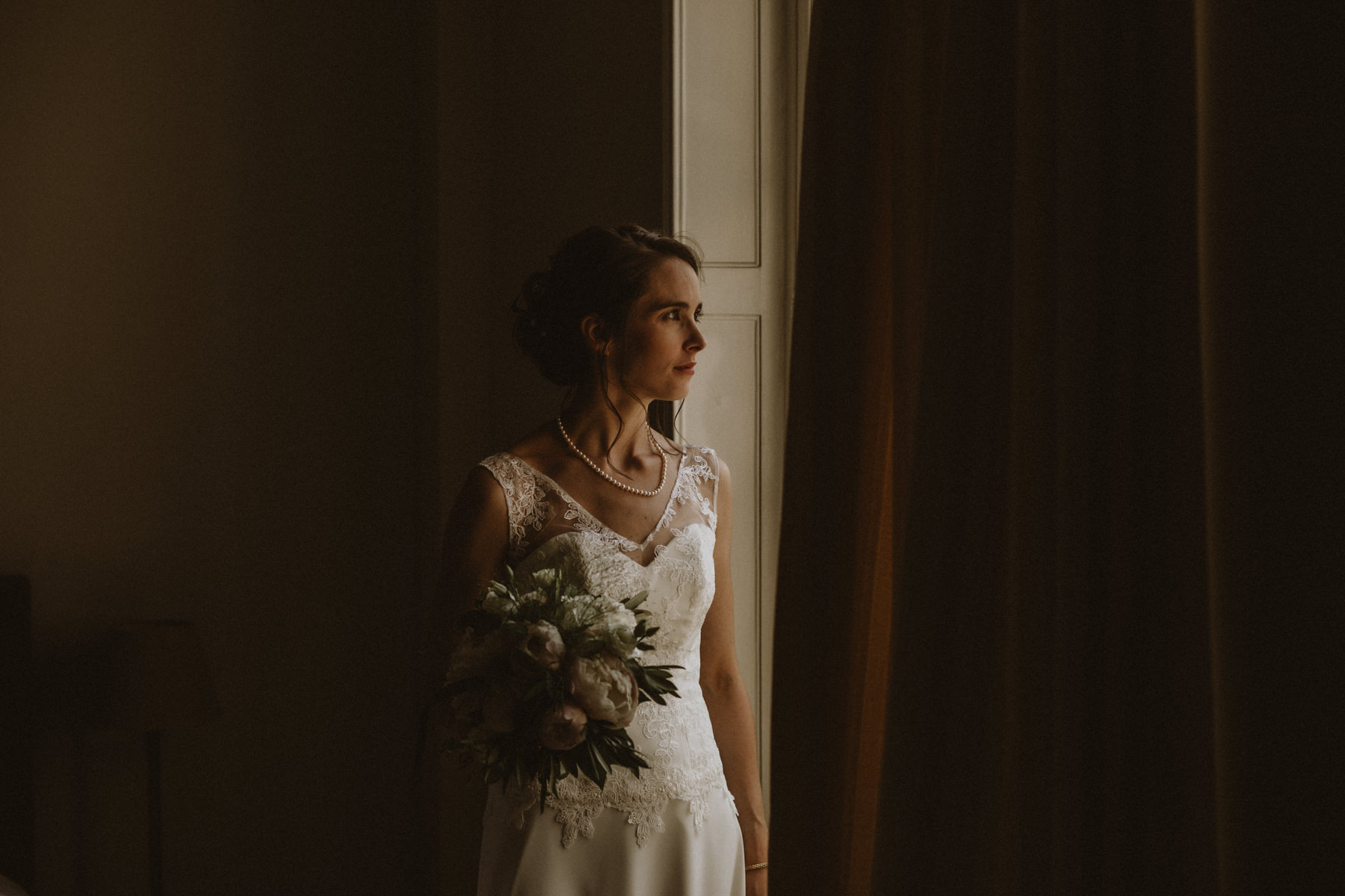 mariée qui regarde par le fenêtre avec son bouquet à la main à la maison de myon